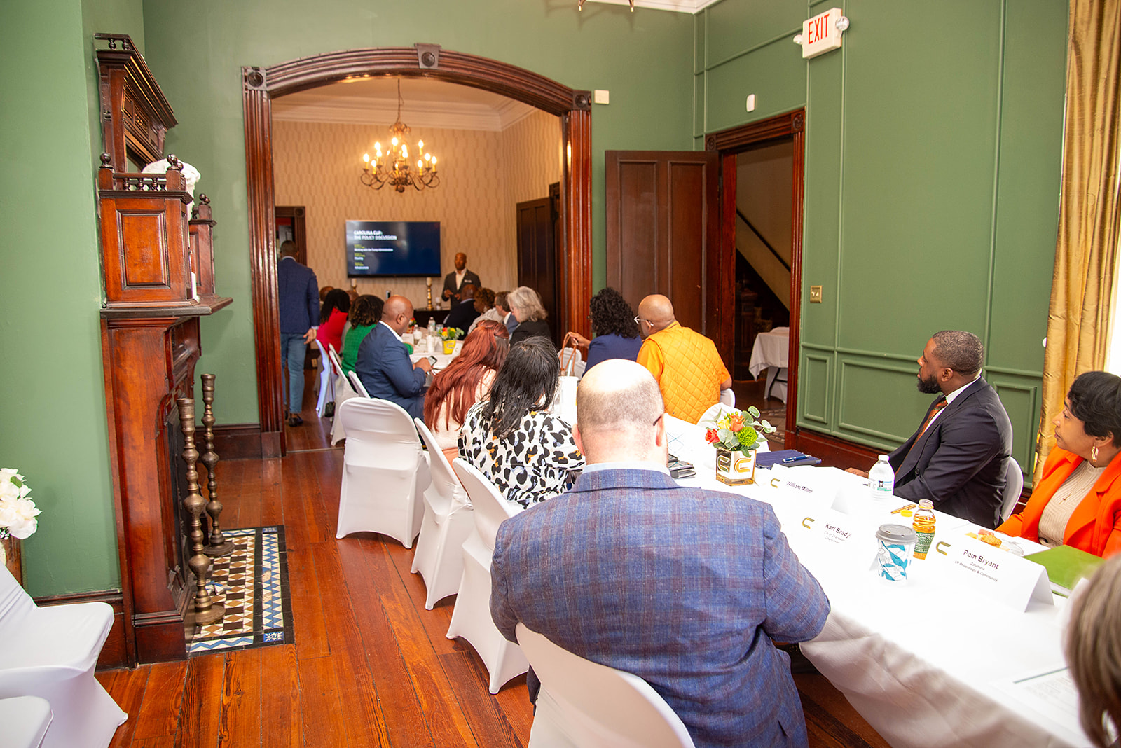 A group of professionals seated around a long conference table in a historic room with green walls and wooden floors, attending a policy discussion. A speaker is presenting in an adjacent room with a screen displaying a presentation.