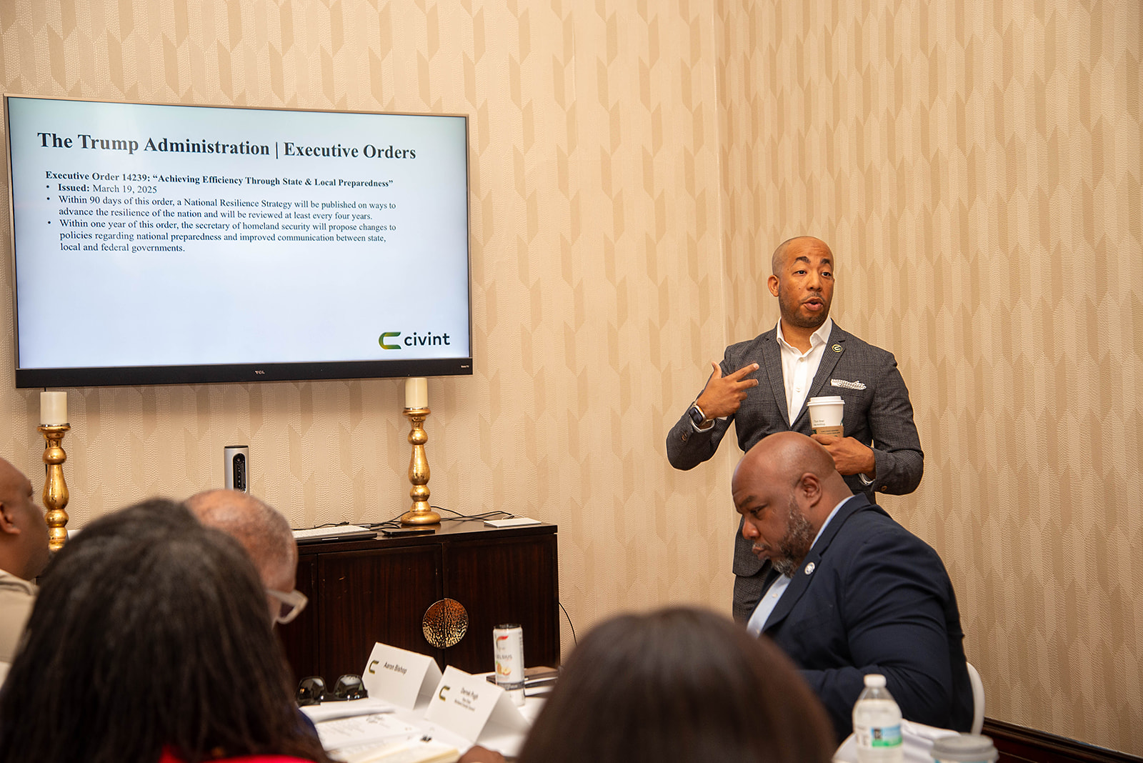 Sam Johnson, in a gray checked suit jacket, holding a coffee cup, speaks during a policy discussion in front of a screen displaying a presentation.