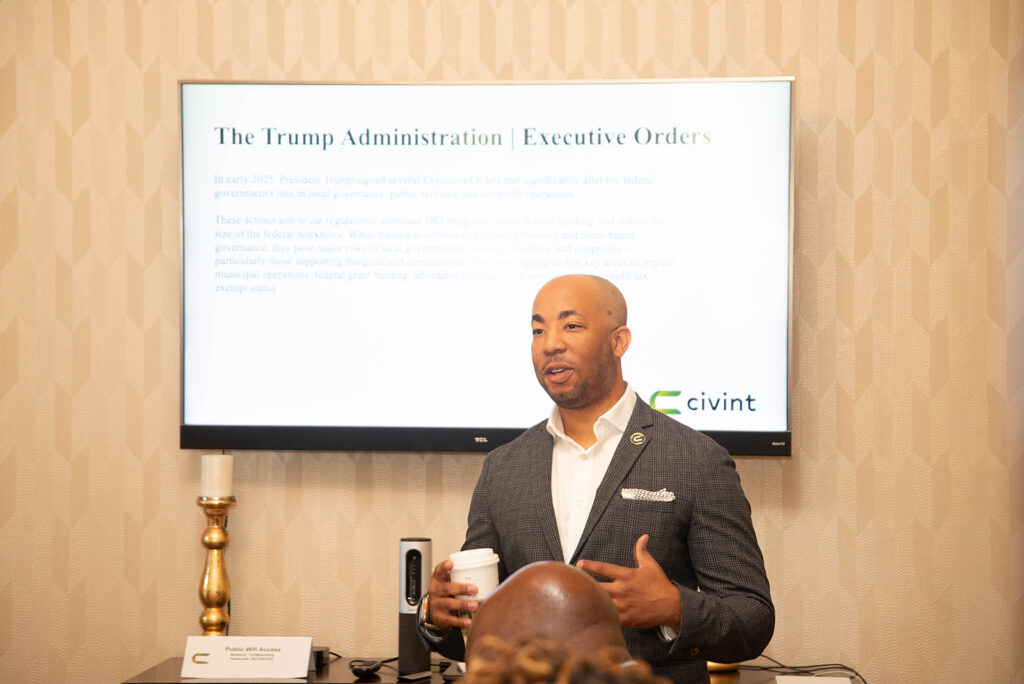 Sam Johnson, in a gray checked suit jacket, holding a coffee cup, speaks during a policy discussion in front of a screen displaying a presentation.